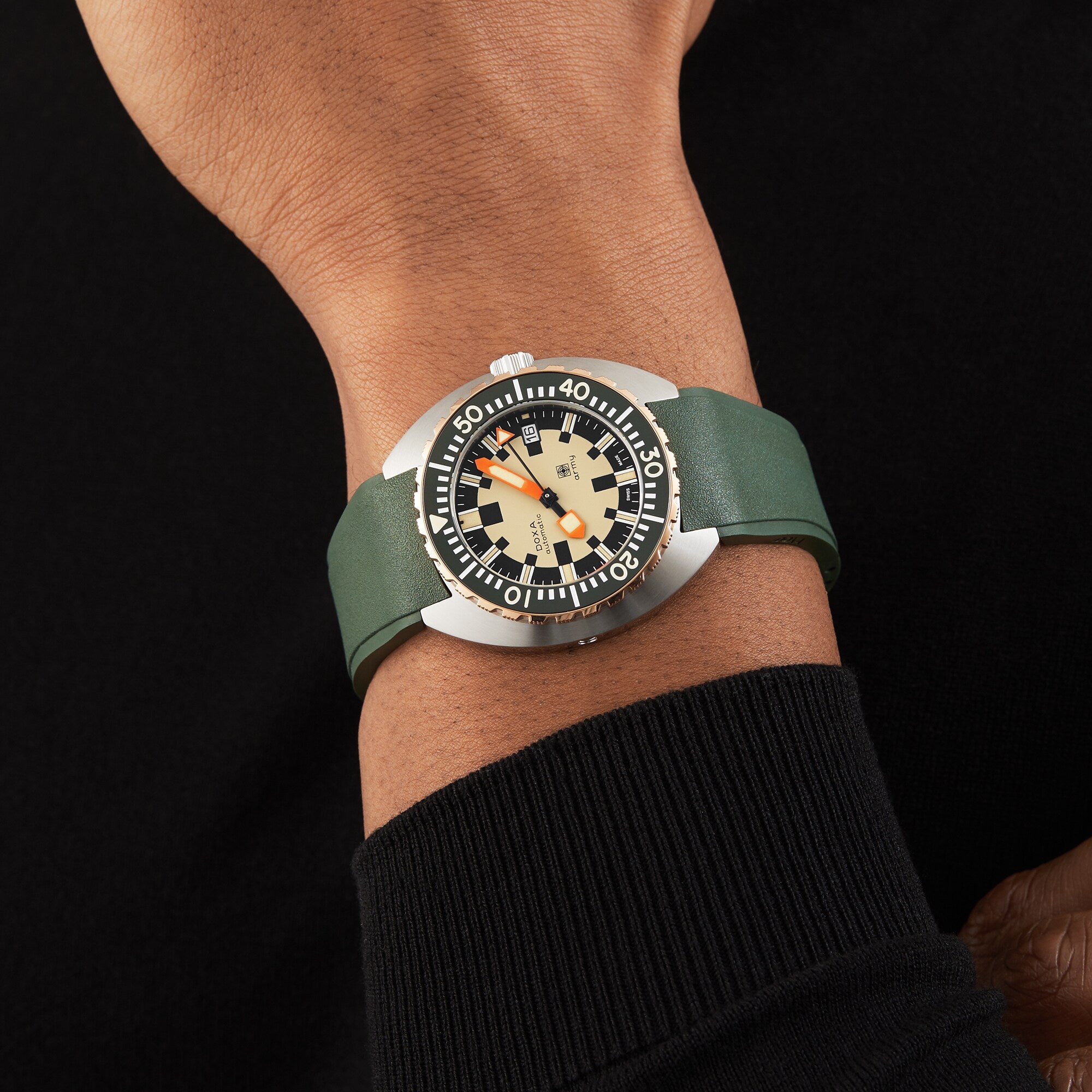 A person wears a DOXA Army Bronze by DOXA with a green strap, beige and black dial, orange accents, and a stainless steel uni-directional rotating bezel, paired with a black long-sleeve shirt.