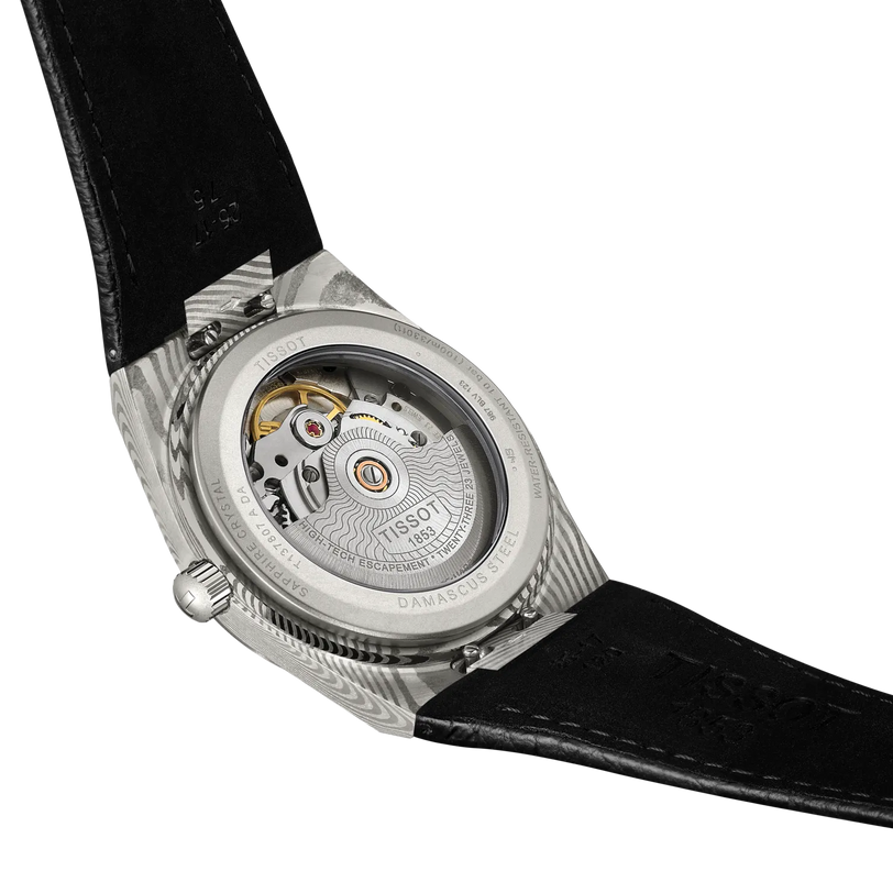 The image displays the back of a Tissot PRX 38 watch with a transparent case, showcasing its intricate automatic movement. It features a black leather strap and an engraved stainless steel case.