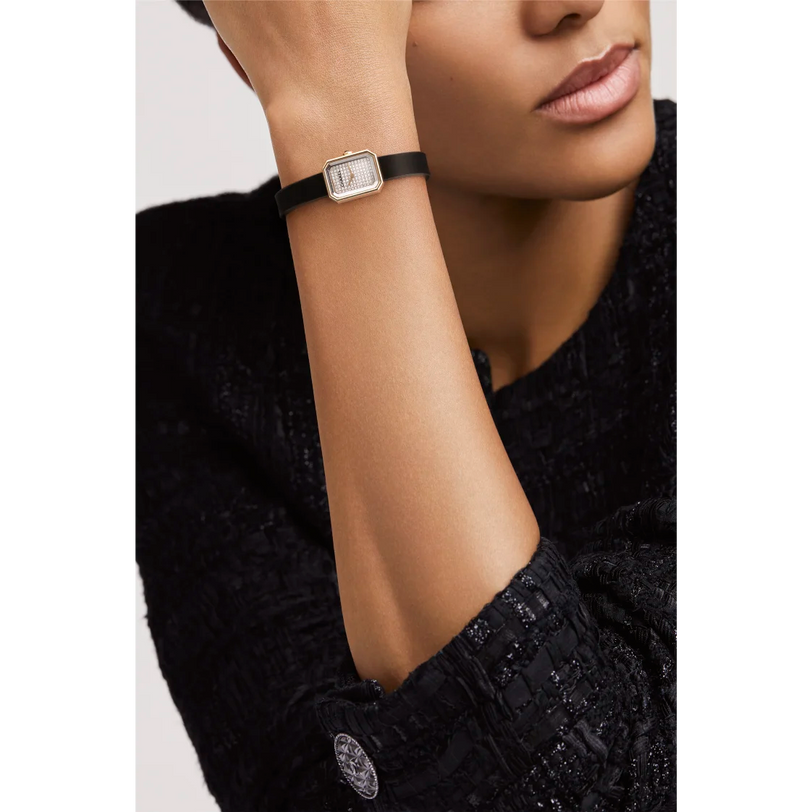 A person in a textured black jacket rests their chin on their hand, displaying the Chanel Première Ribbon watch with a diamond dial, black strap, and gold-tone case.