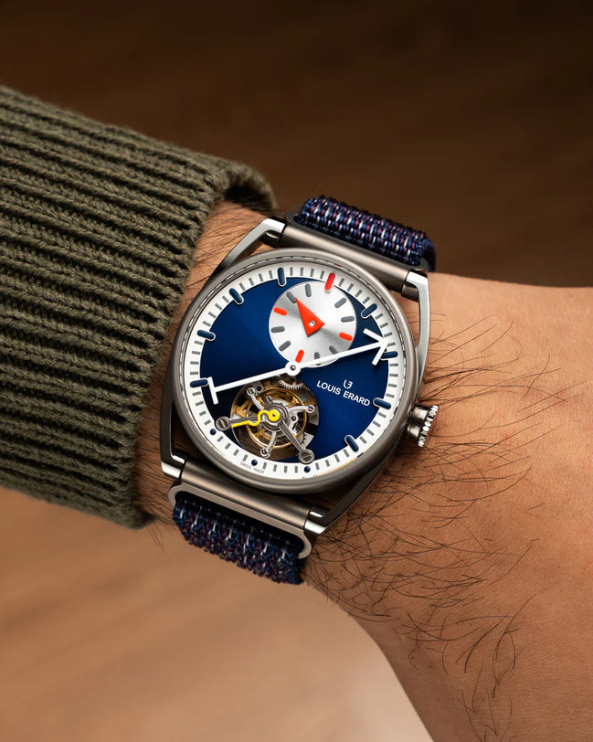 Stainless steel watch with blue dial, white subdial, red accents, visible tourbillon, and navy fabric strap worn on wrist.