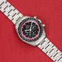 An OMEGA Speedmaster Tintin watch with a black dial and silver case displayed on a red textured background.