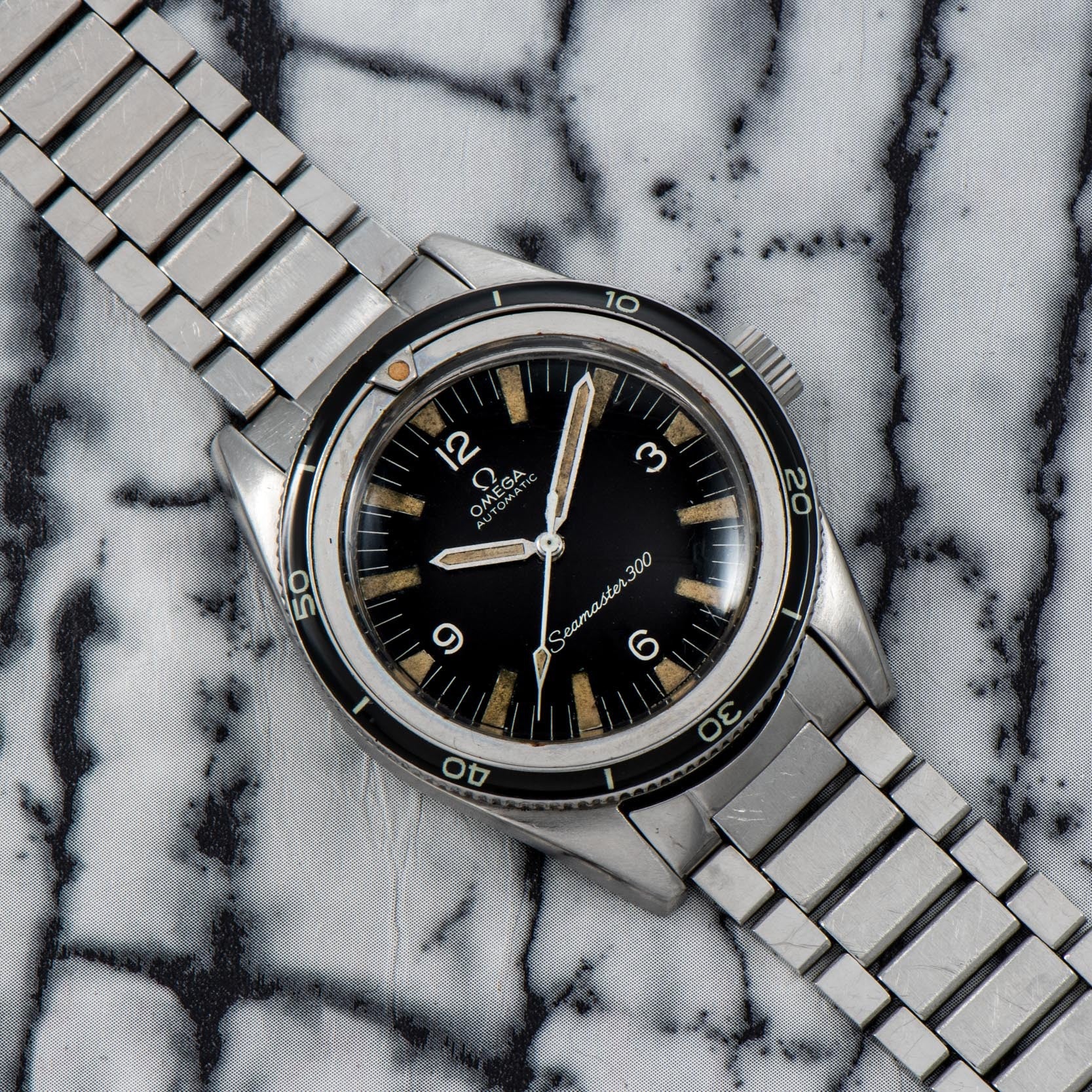 An OMEGA Seamaster 300 watch with a metal band displayed on a marble-patterned surface.