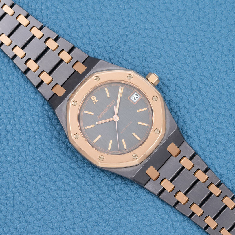 The Audemars Piguet Royal Oak Tantalum luxury wristwatch, featuring a gold bezel and black strap, is displayed on a textured blue surface.