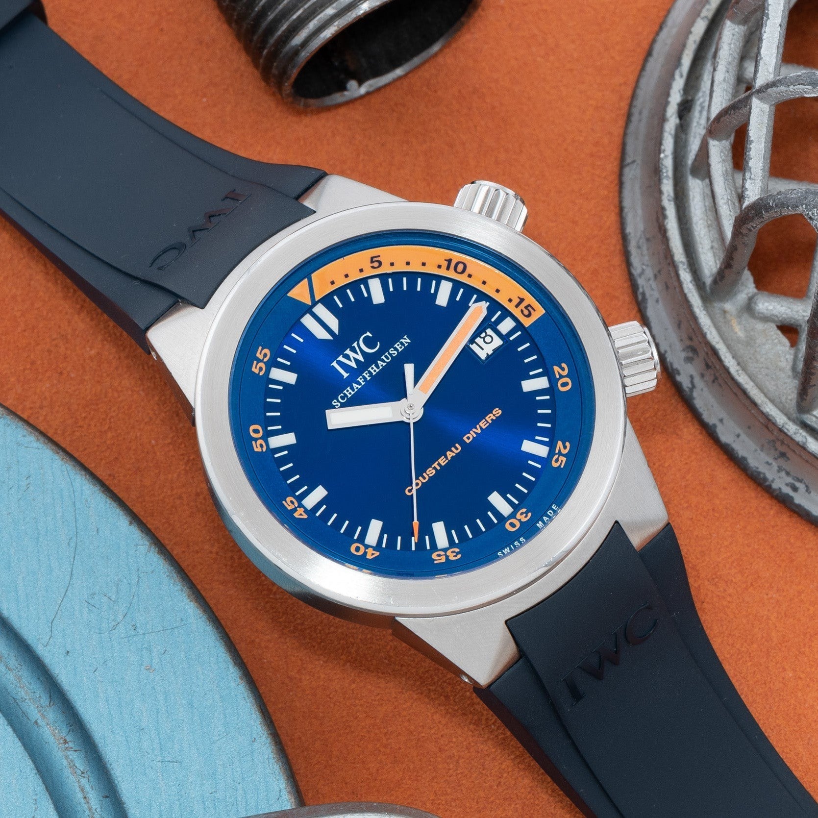 An IWC Schaffhausen Aquatimer Cousteau Divers Limited Edition with a blue dial and black strap is displayed on an orange surface beside metal objects.