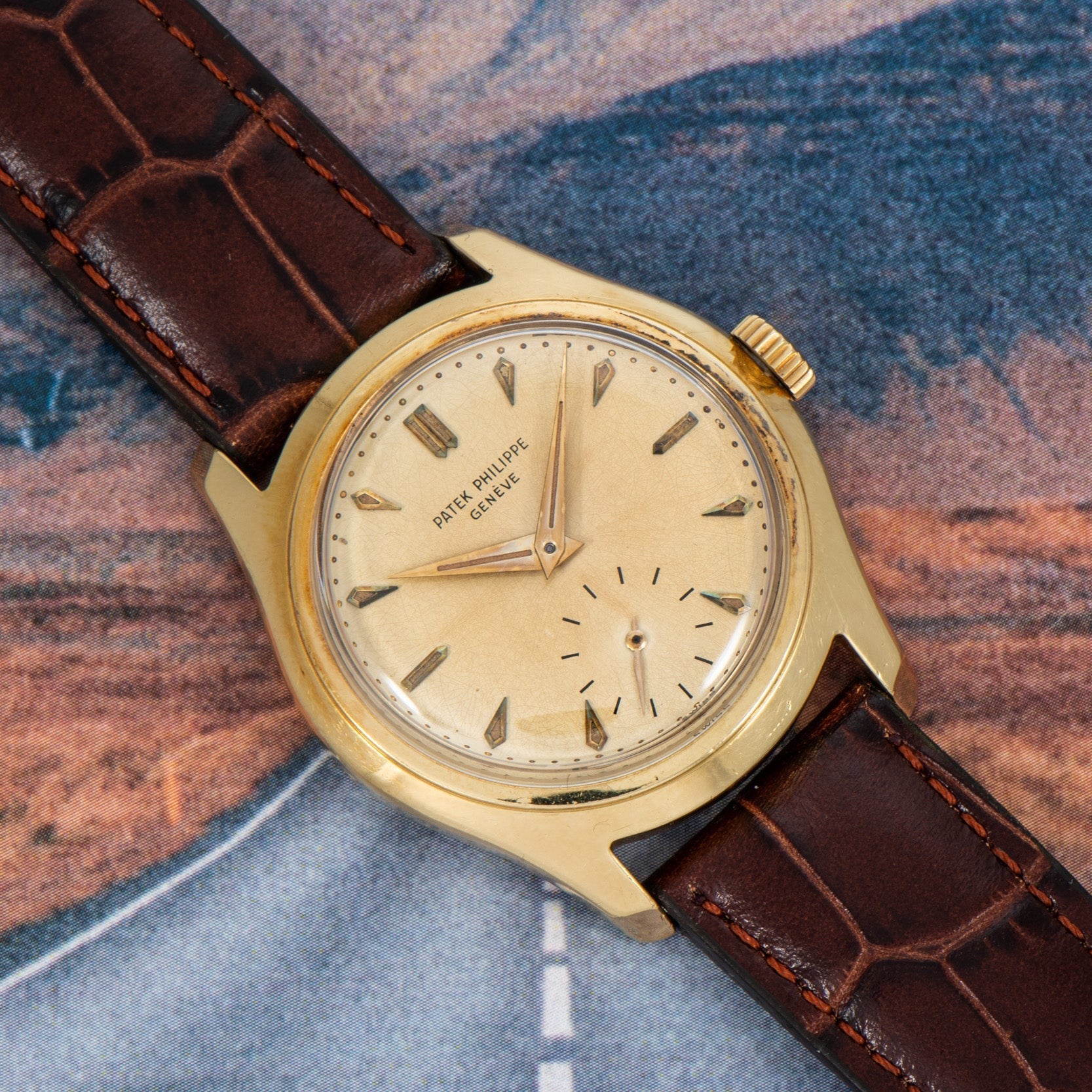 A Patek Philippe Calatrava gold wristwatch with a brown leather strap is displayed against a blurred road background.