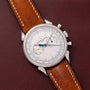 The Vacheron Constantin Historiques Cornes De Vache Chronograph, featuring a silver case and brown leather strap, rests elegantly on a dark brown leather surface.