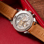 The Vacheron Constantin Historiques Cornes De Vache Chronograph, featuring a brown leather strap and an intricate exhibition caseback, is displayed in a red case.