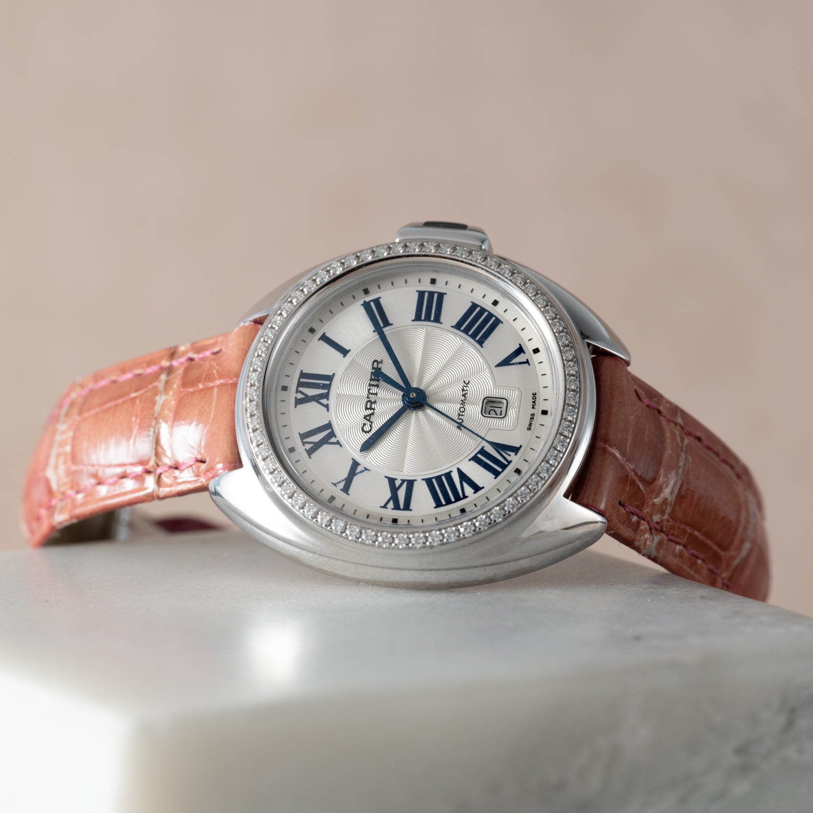 The Cle de Diamond by Cartier, featuring a pink leather strap and blue Roman numerals, rests on a marble surface.