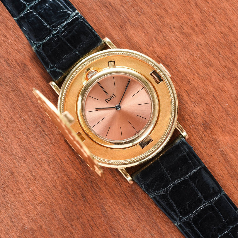 The Piaget 1877 Twenty Dollar Coin Watch features a gold case, pink dial, black leather strap, and an open protective cover.