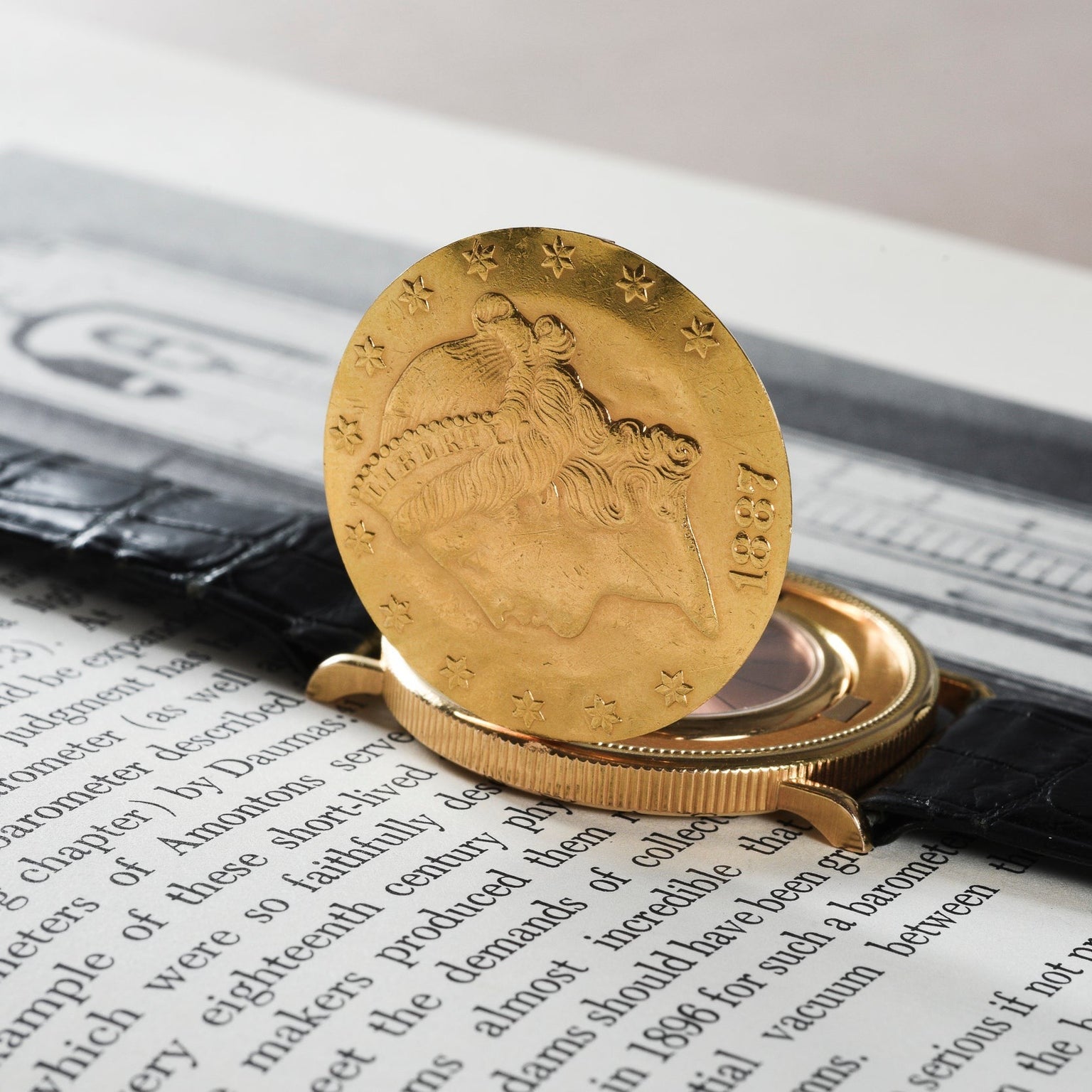A Piaget 1877 Twenty Dollar Coin Watch with a black leather strap rests on an open book.