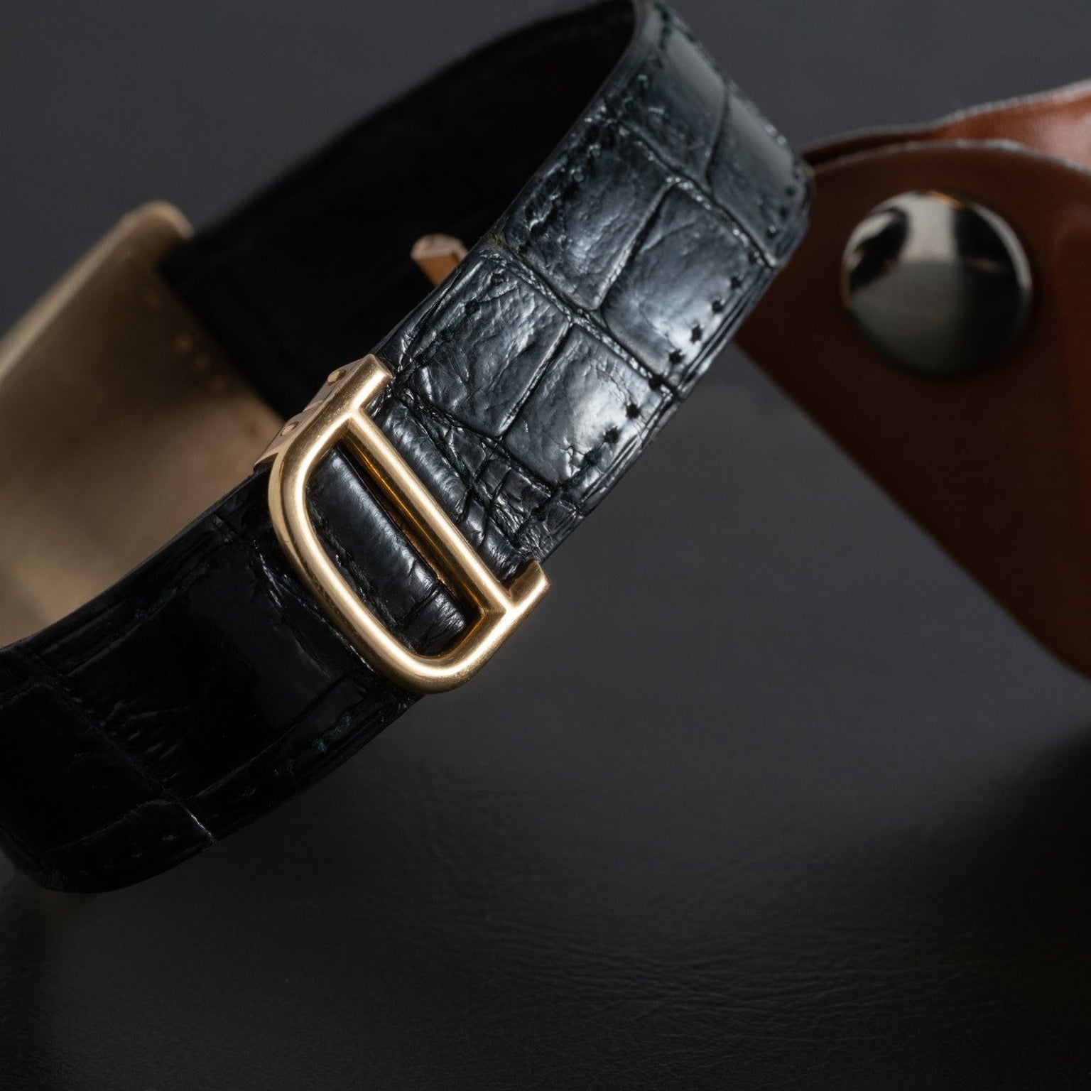 Close-up of the Cartier Tank Cintrée Jumbo black leather watch strap with a gold buckle, placed beside a brown leather item on a dark surface.