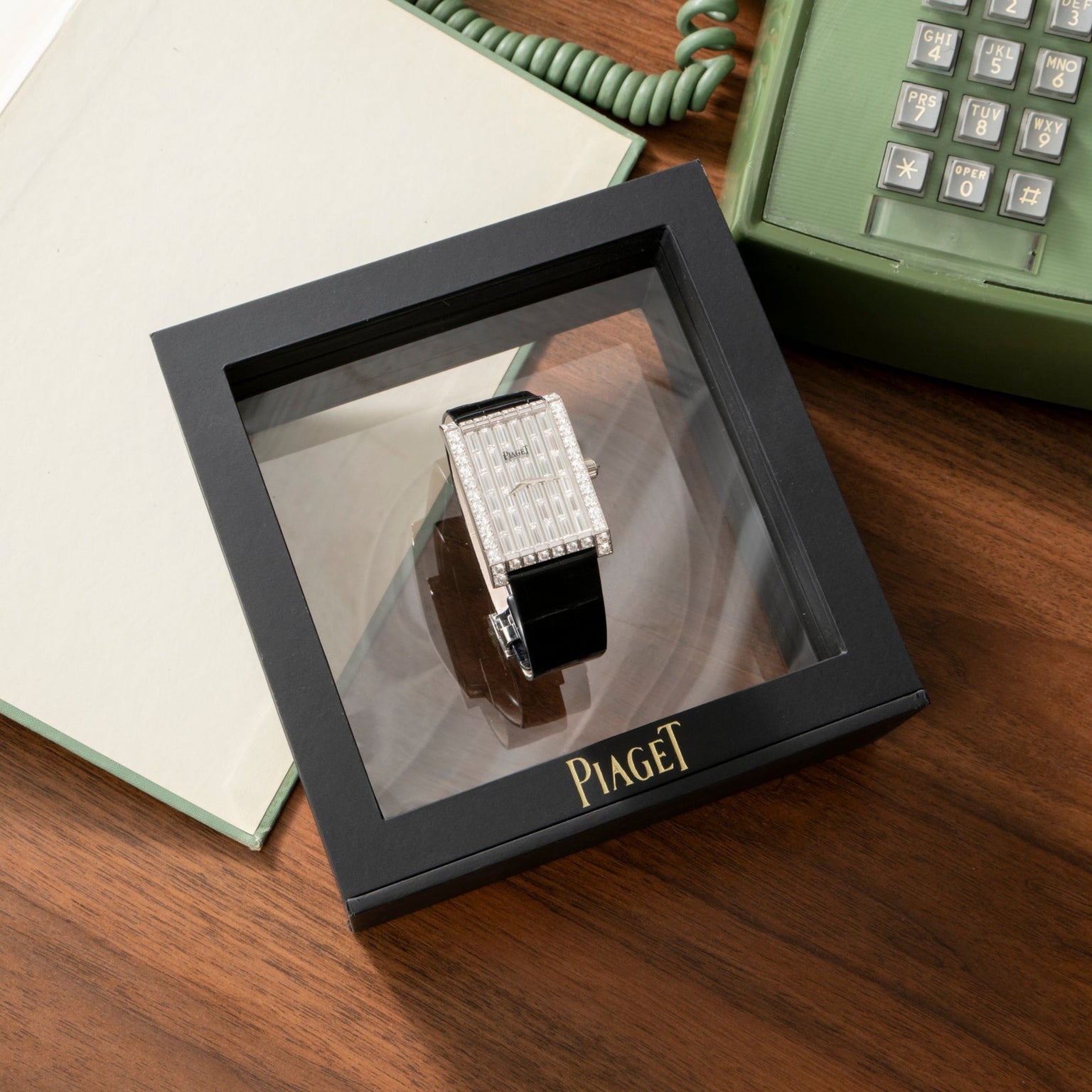 A Piaget Limelight Baguette Diamond Dress Watch with a jeweled rectangular face, displayed in a black box on a wooden desk.