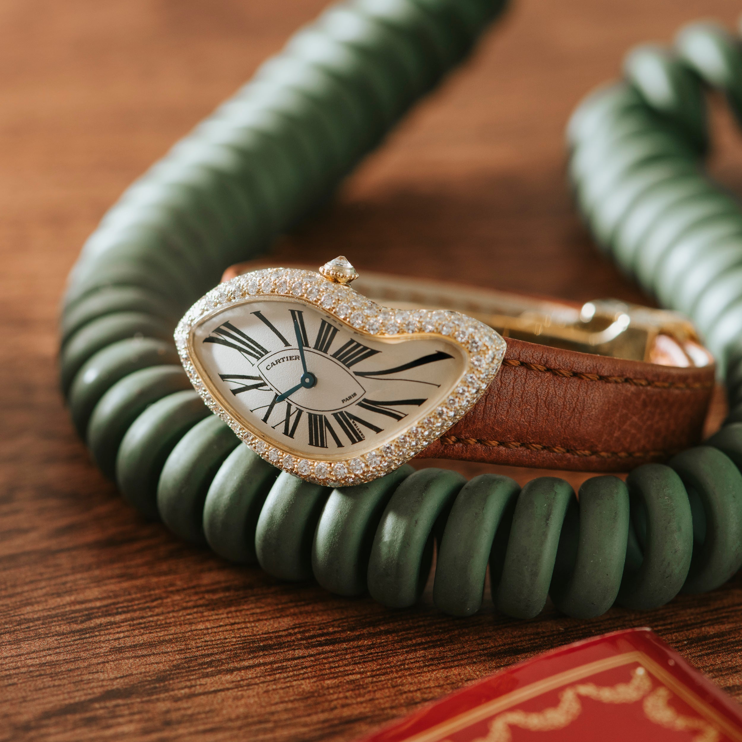 A Cartier Crash Diamonds watch with a brown strap lies on a wooden surface, encircled by a green coiled cord.