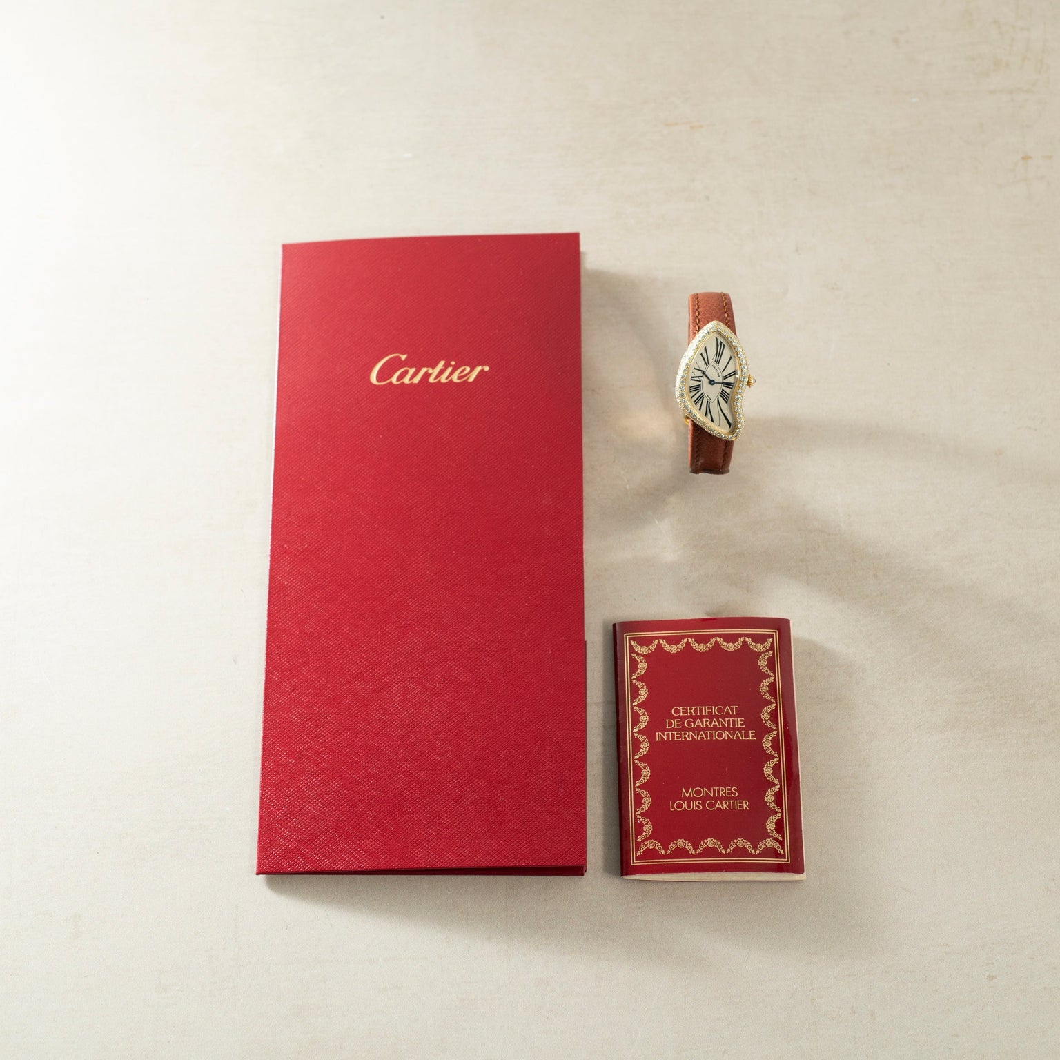 A Cartier Crash Diamonds watch with a brown strap, red warranty card, and red Cartier box displayed on a light surface.