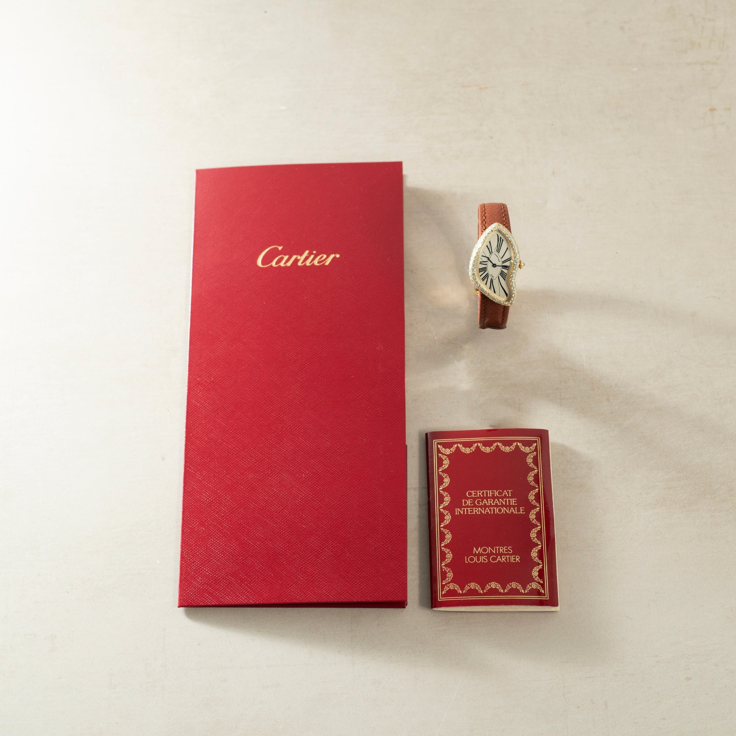 A Cartier Crash Diamonds watch with a brown strap, red warranty card, and red Cartier box displayed on a light surface.