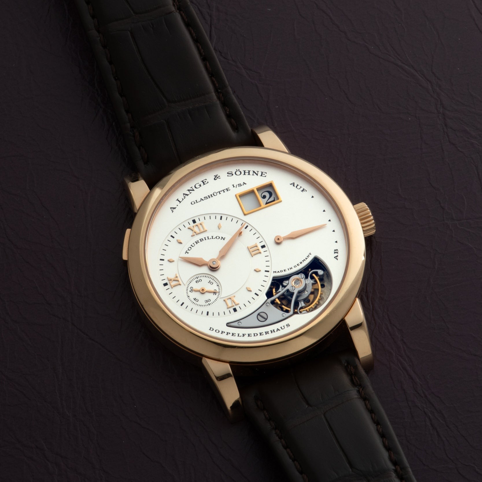 The Lange & Söhne Lange 1 Tourbillon features a gold case, leather strap, and visible tourbillon, elegantly displayed on a dark surface.