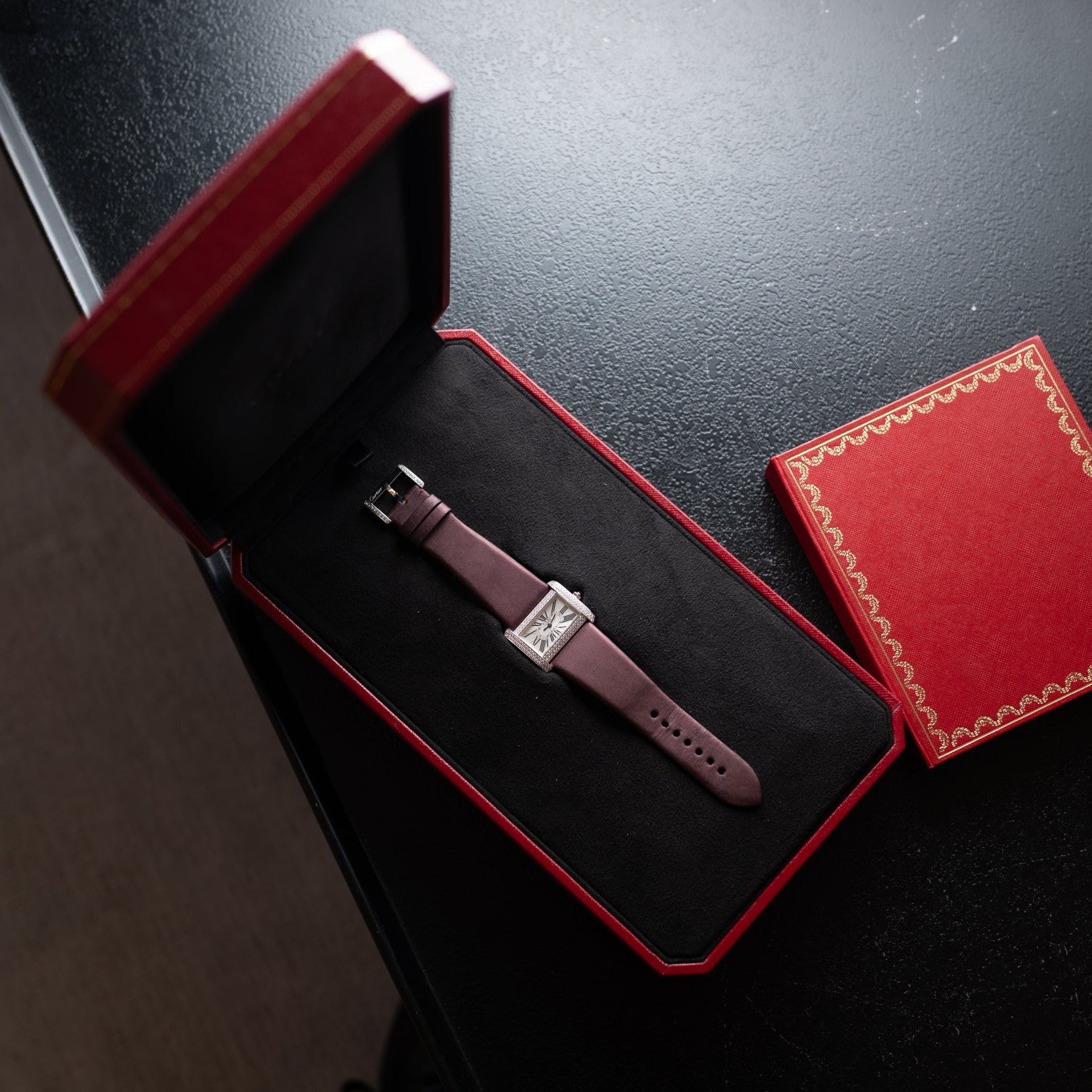 A Cartier Tank Divan wristwatch in brown leather, placed in a red gift box on a black table.