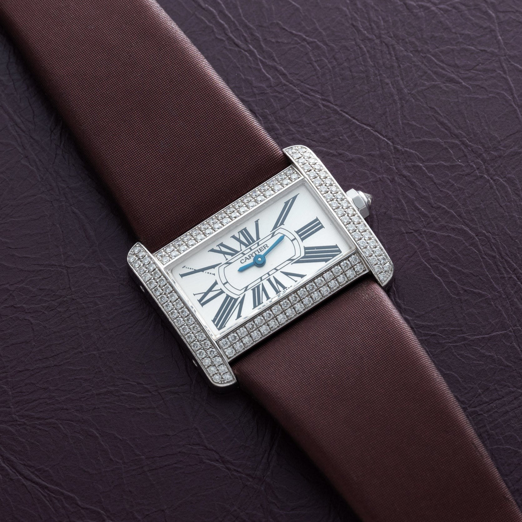The Cartier Tank Divan, a rectangular watch with diamond accents and a brown strap, rests on a textured dark surface.