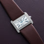 The Cartier Tank Divan, a rectangular watch with diamond accents and a brown strap, rests on a textured dark surface.