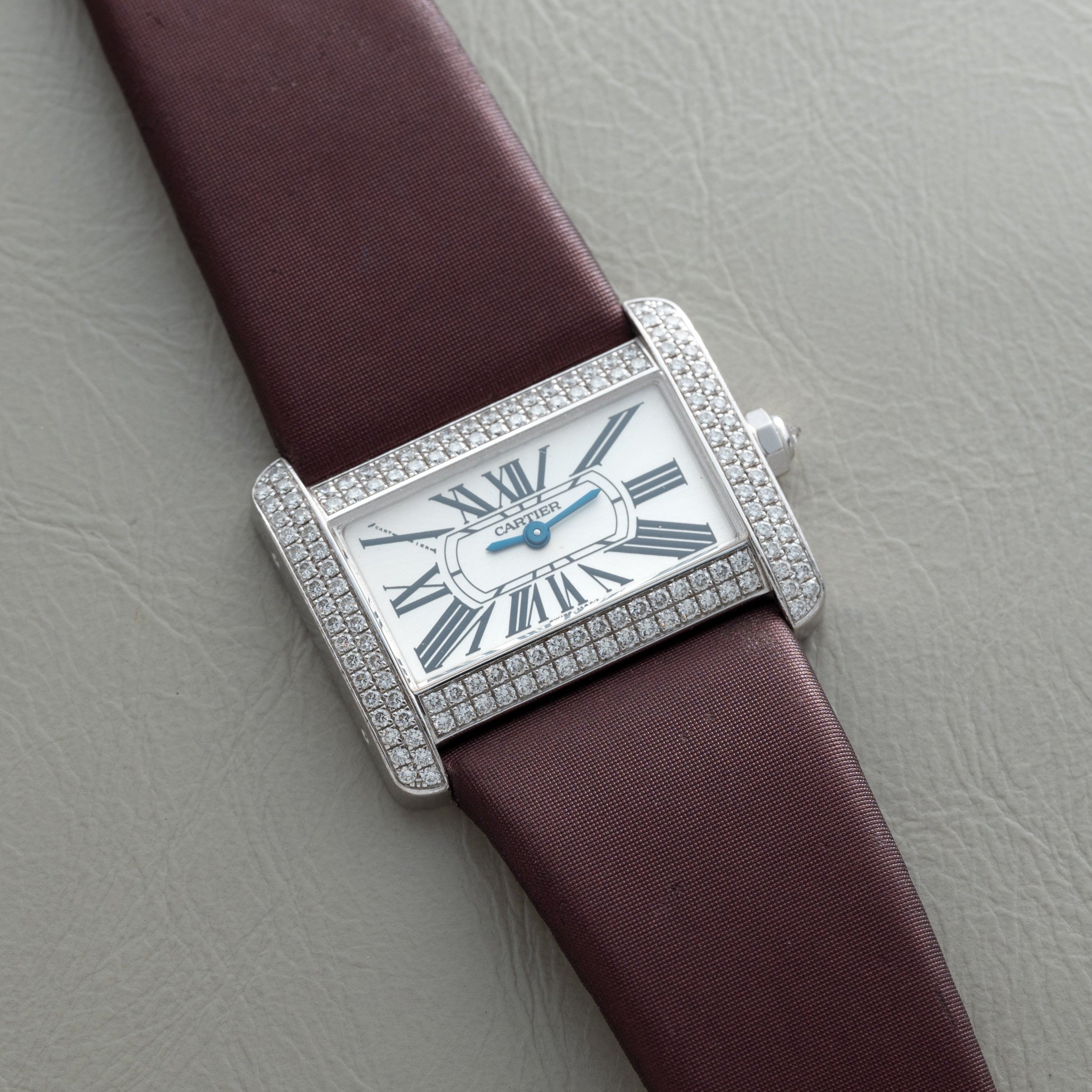 A Cartier Tank Divan watch with a diamond bezel, Roman numerals, and a brown strap is displayed on a gray surface.