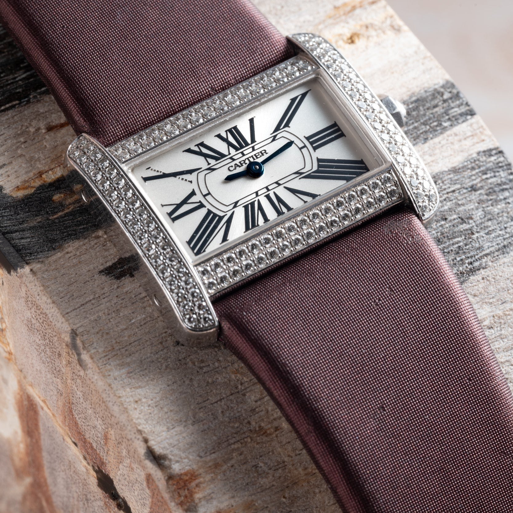 A Cartier Tank Divan watch with a diamond bezel, Roman numerals, and a purple strap rests on a wooden surface.