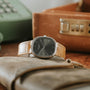 A Patek Philippe Ellipse wristwatch with a tan leather strap and black face rests on a brown leather pouch near a briefcase.