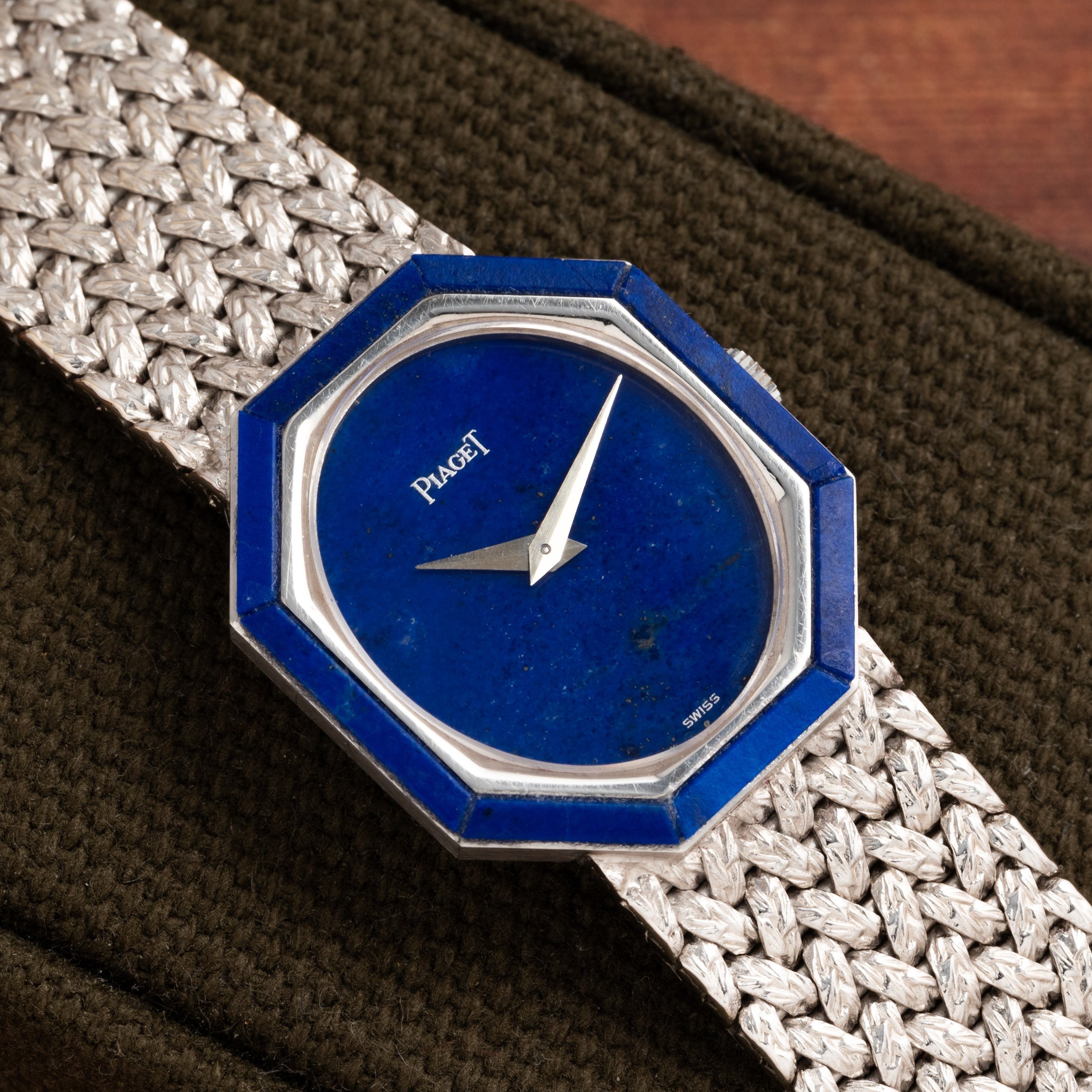 The Piaget Lapis Lazuli White Gold Dress Watch, featuring a blue octagonal face and woven metal strap, rests elegantly on a textured dark surface.