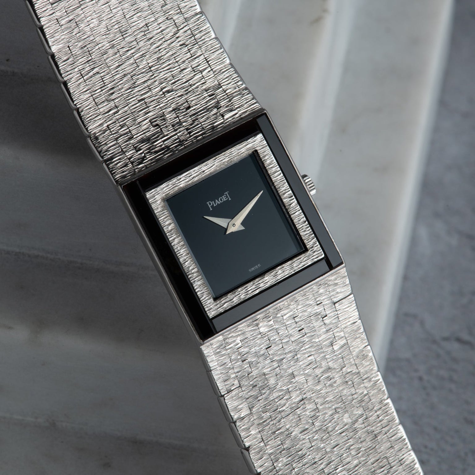 A Piaget White Gold Onyx Dress Watch with a textured band and rectangular black dial is displayed on a gray marble surface.