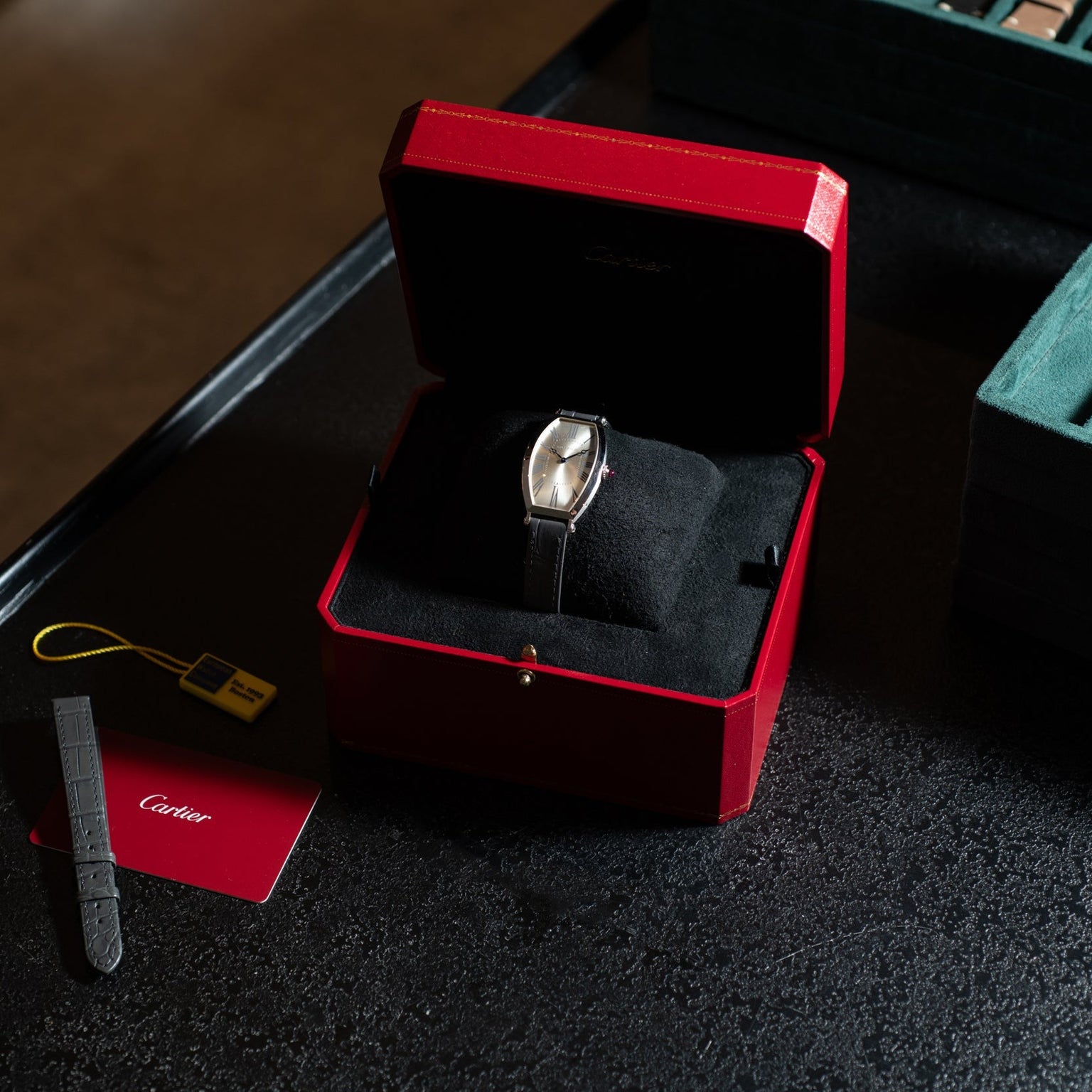 A Cartier Tonneau Platinum wristwatch in a red box, accompanied by a card, tag, and extra strap on a dark surface.