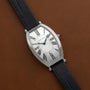 A Cartier Tonneau Platinum watch with a silver face, Roman numerals, blue hands, and a black leather strap rests on a brown surface.