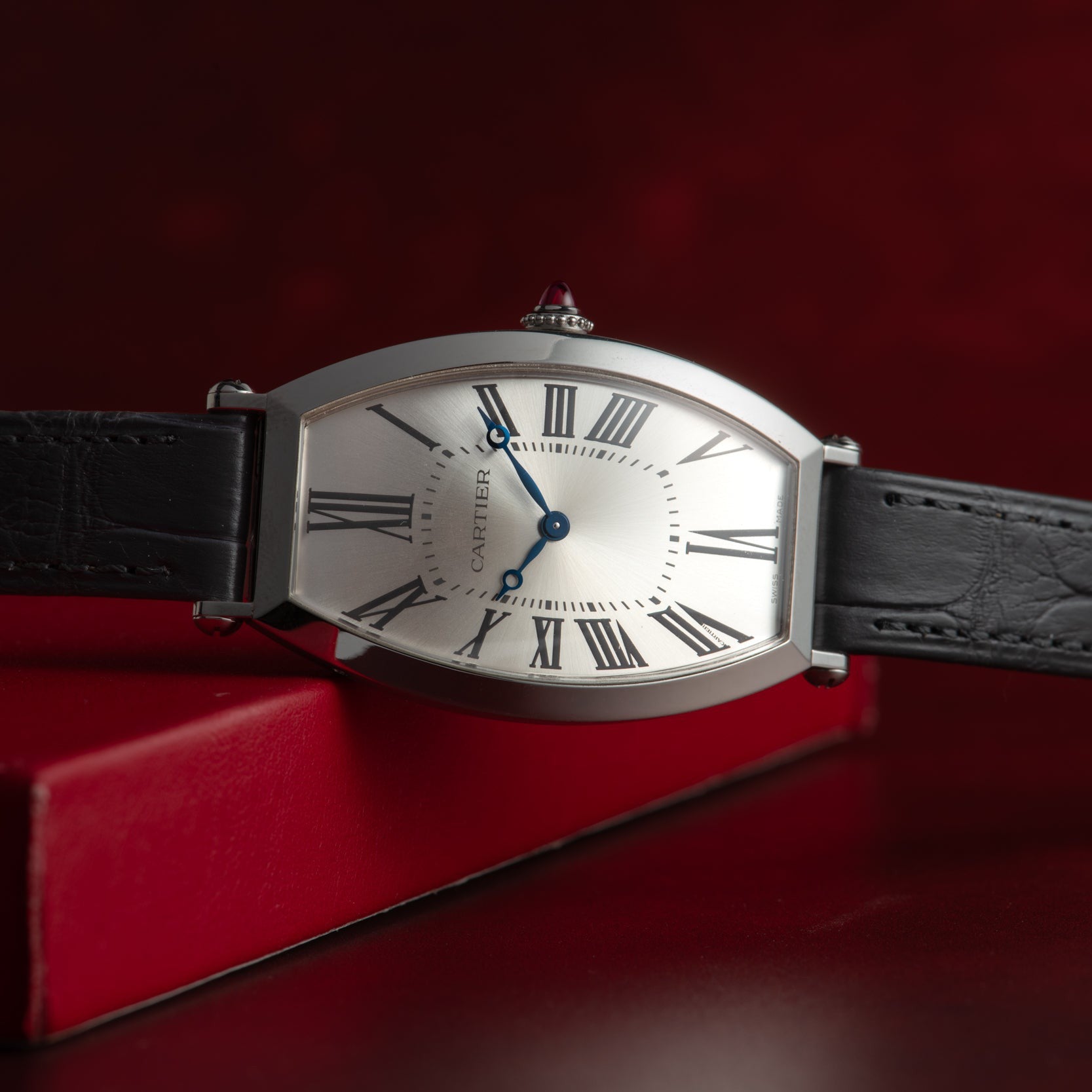 The Cartier Tonneau Platinum wristwatch, featuring a black leather strap and Roman numerals, is displayed against a red background.