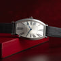 The Cartier Tonneau Platinum wristwatch, featuring a black leather strap and Roman numerals, is displayed against a red background.