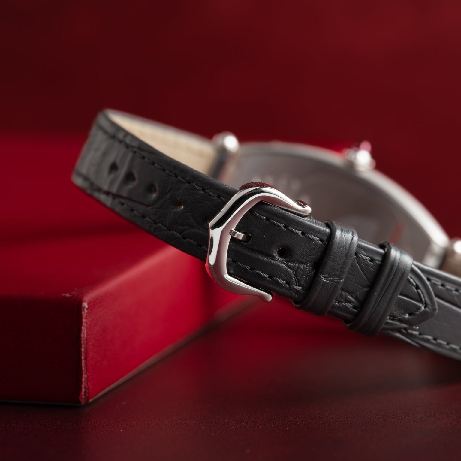 Close-up of Cartiers Tonneau Platinum black leather strap with silver buckle, resting next to a red box on a red background.