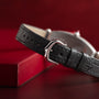 Close-up of Cartiers Tonneau Platinum black leather strap with silver buckle, resting next to a red box on a red background.