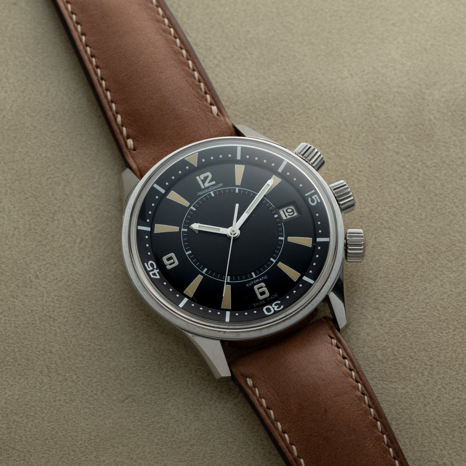 The Jaeger-LeCoultre Tribute to Polaris, a vintage-style watch with a brown leather strap and black dial, rests on a beige surface.