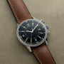 The Jaeger-LeCoultre Tribute to Polaris, a vintage-style watch with a brown leather strap and black dial, rests on a beige surface.