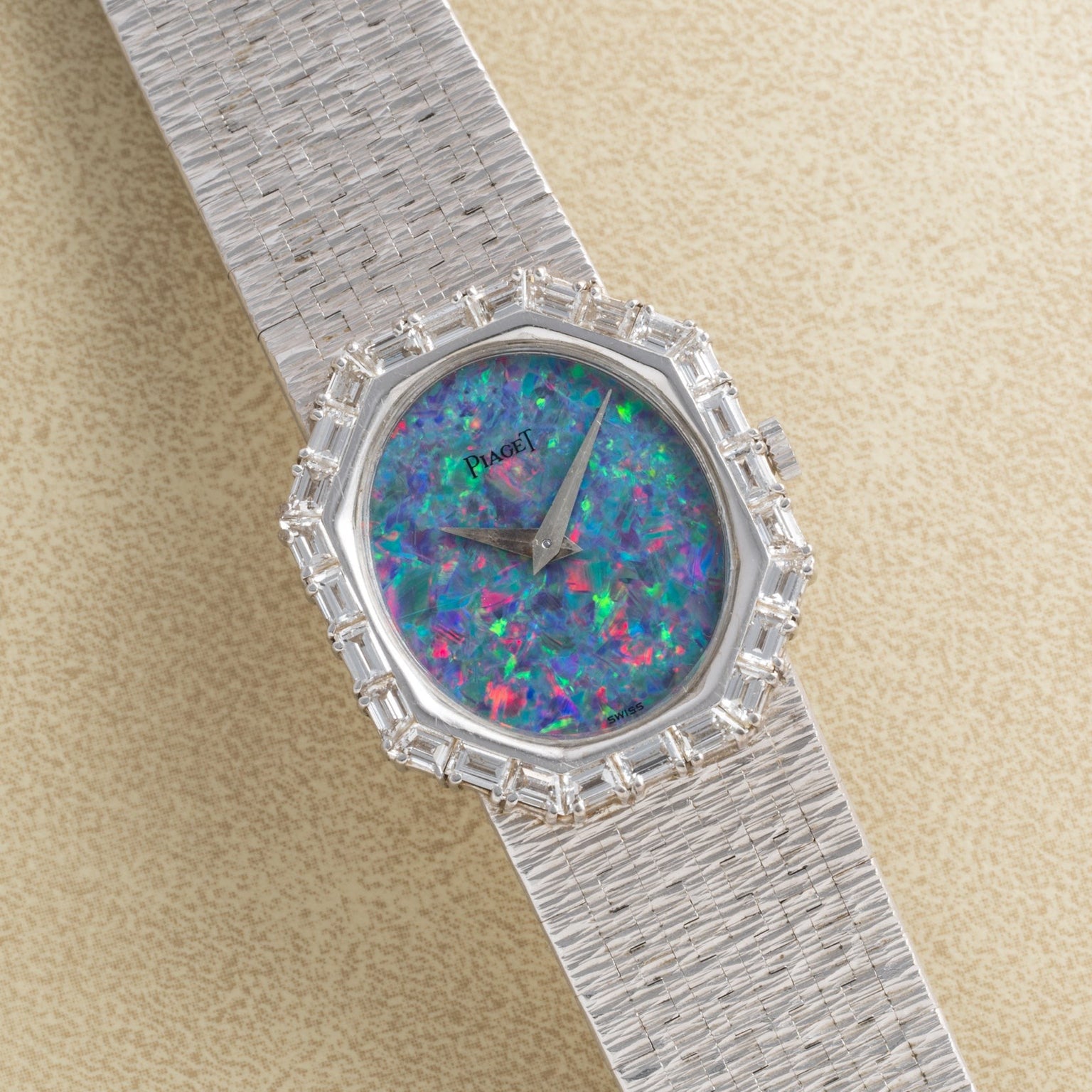 The Piaget Opal White Gold Dress Watch features a colorful opal face, diamond bezel, and textured band, elegantly displayed at an angle on a beige surface.