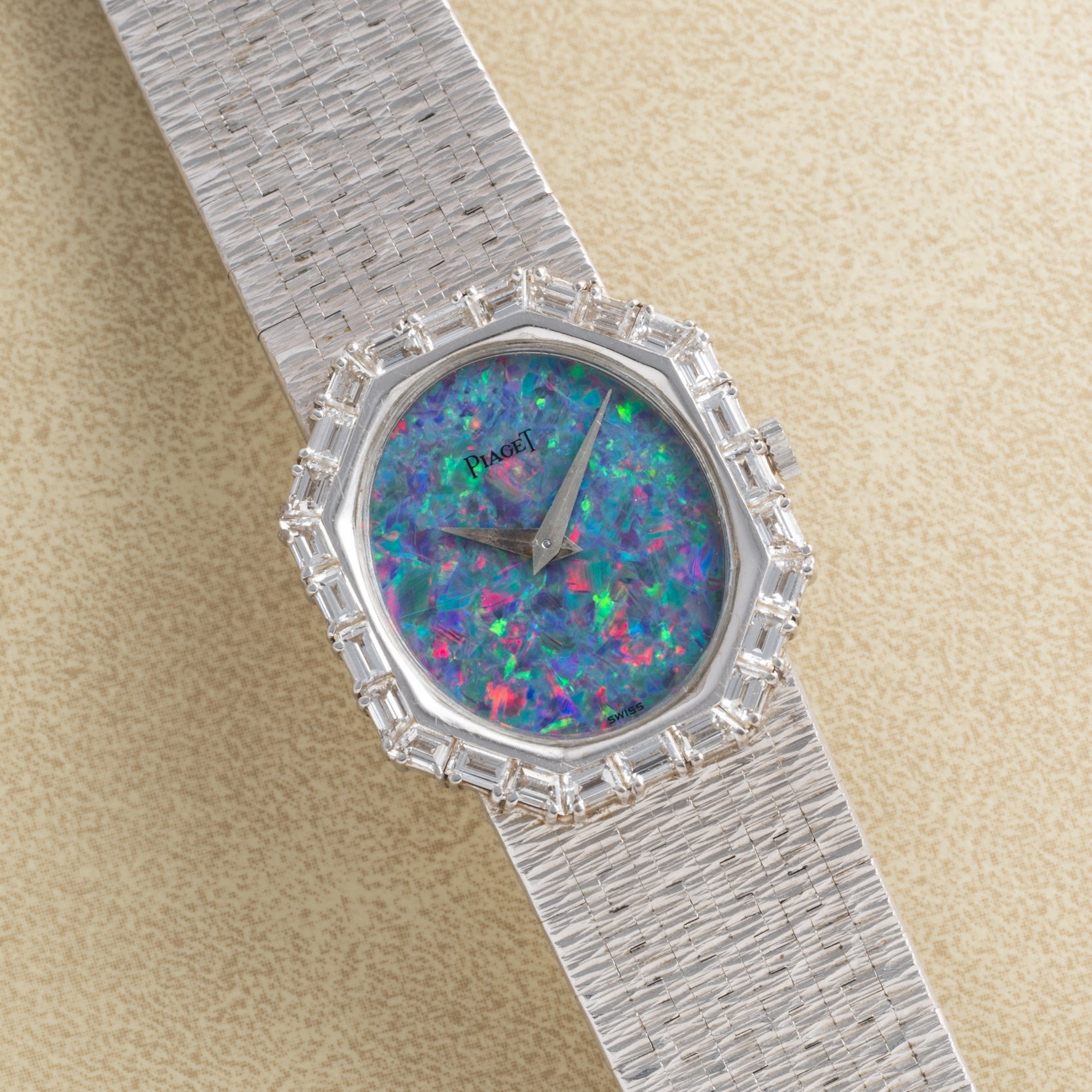 The Piaget Opal White Gold Dress Watch features a colorful opal face, diamond bezel, and textured band, elegantly displayed at an angle on a beige surface.