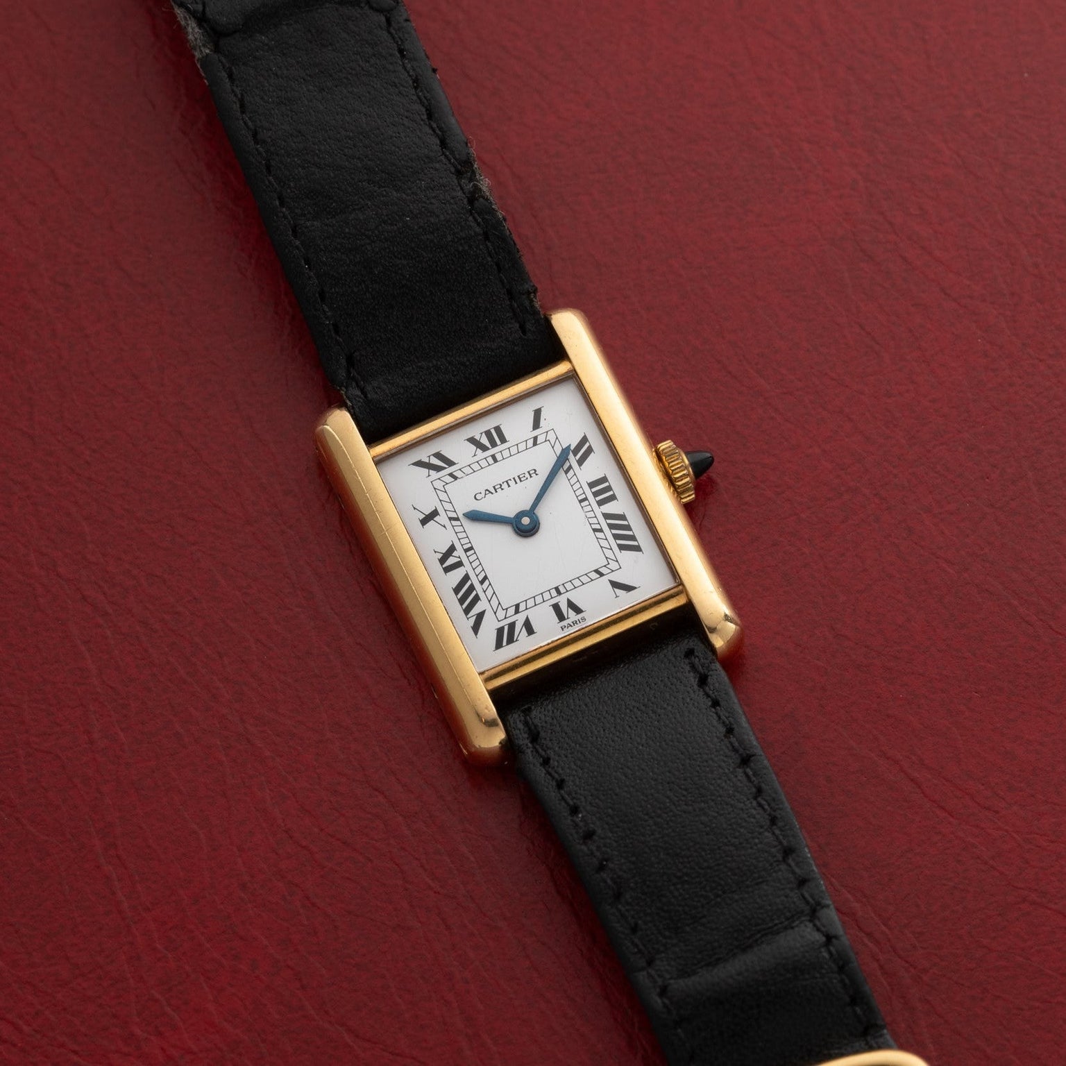 Cartier Tank Piccolo watch featuring a white dial with black Roman numerals and a black leather strap, displayed on a red surface.
