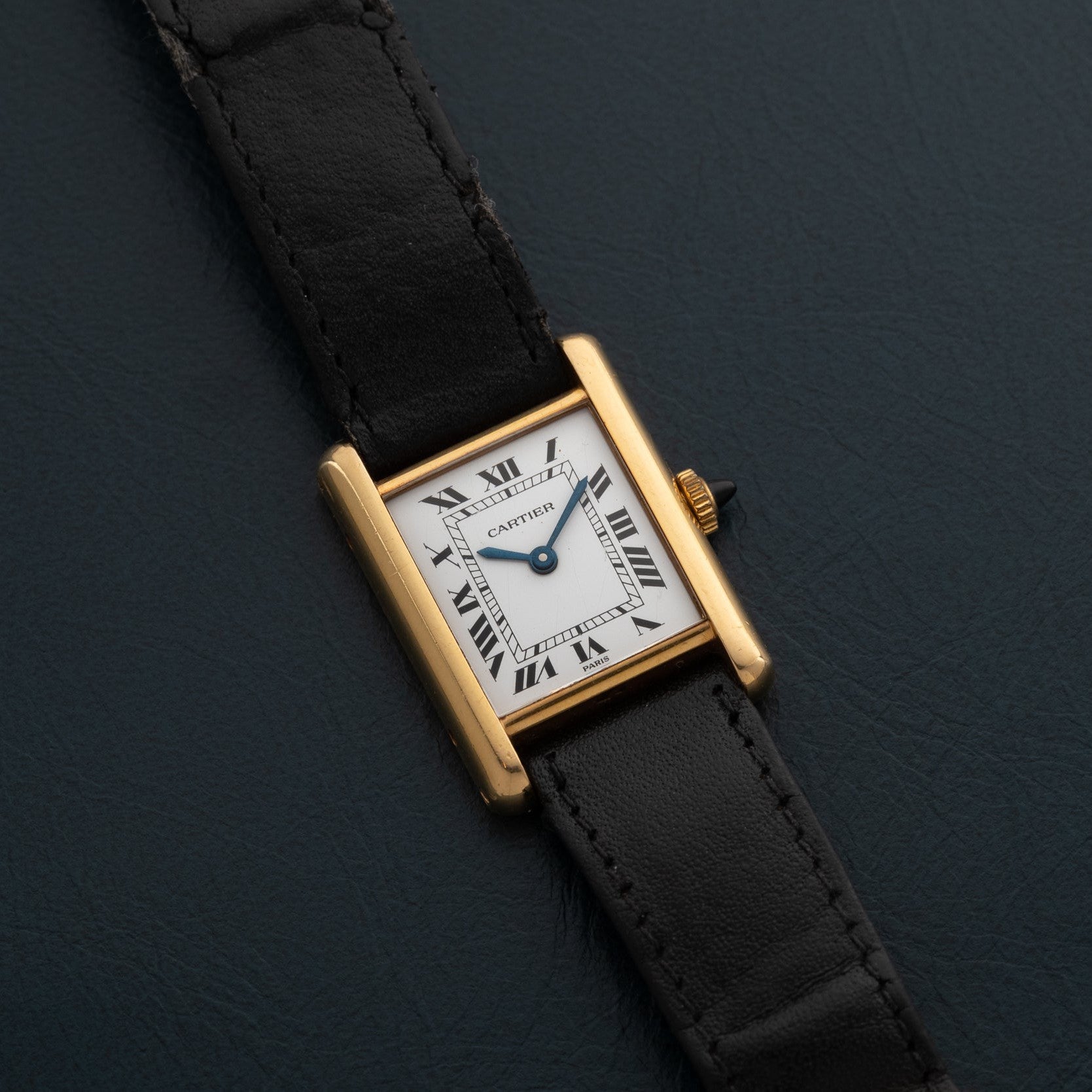 A Cartier Tank Piccolo gold watch with a black leather strap and Roman numeral dial rests on a dark surface.