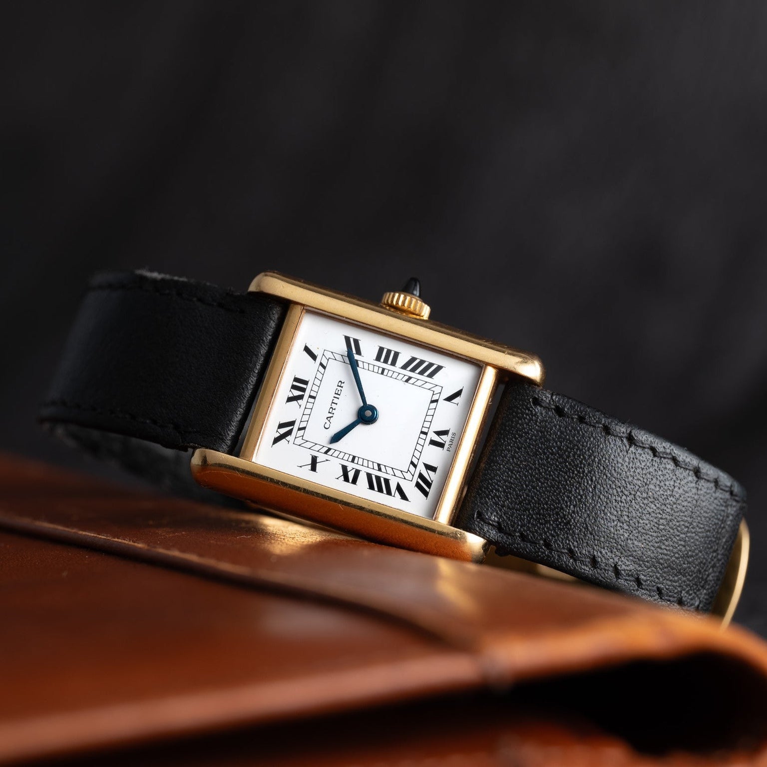 The Cartier Tank Piccolo features a gold rectangular case, black leather strap, and Roman numerals, displayed on a brown leather surface.