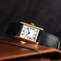 The Cartier Tank Piccolo features a gold rectangular case, black leather strap, and Roman numerals, displayed on a brown leather surface.