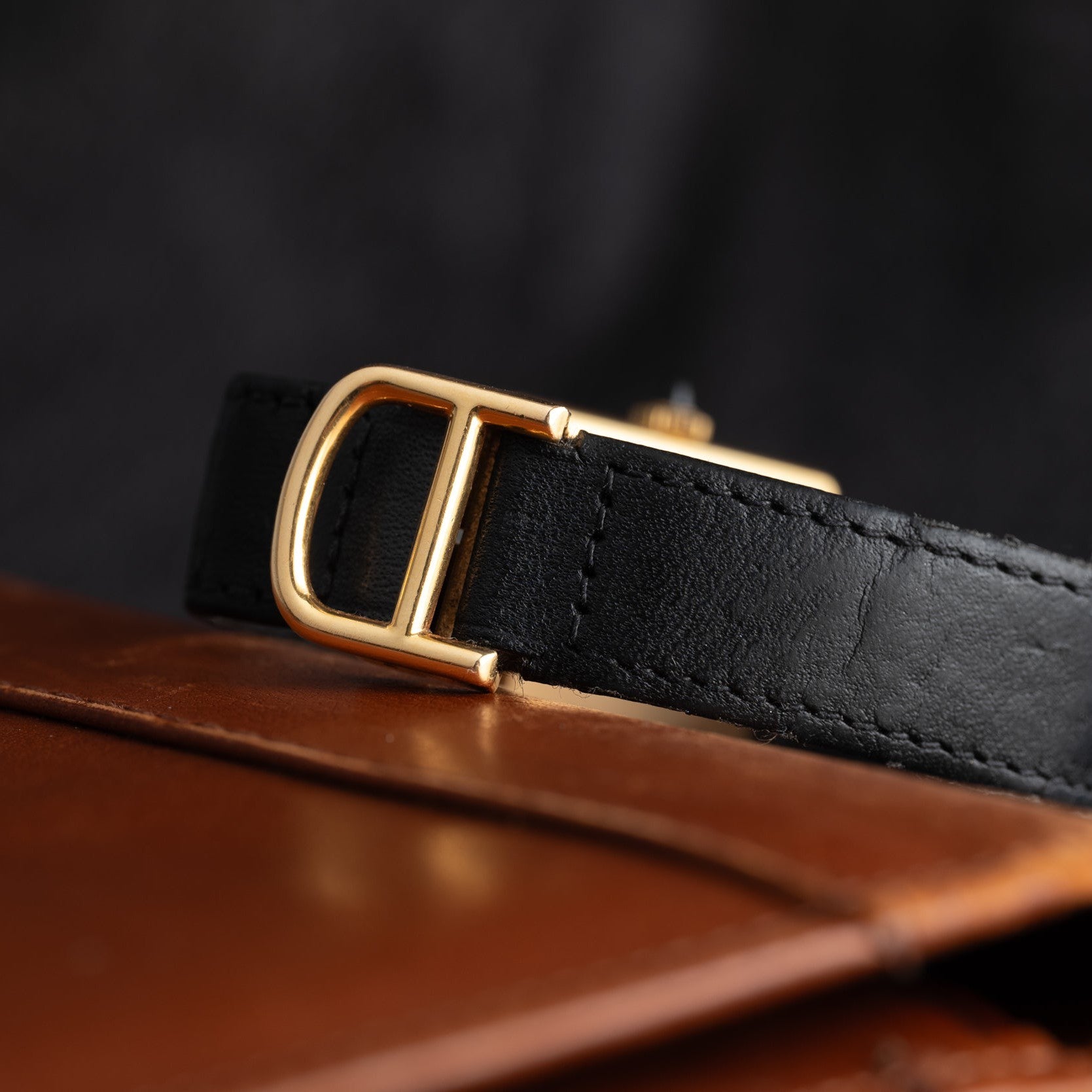 A close-up of the Cartier Tank Piccolo black leather belt with a gold buckle, resting on a brown leather surface.