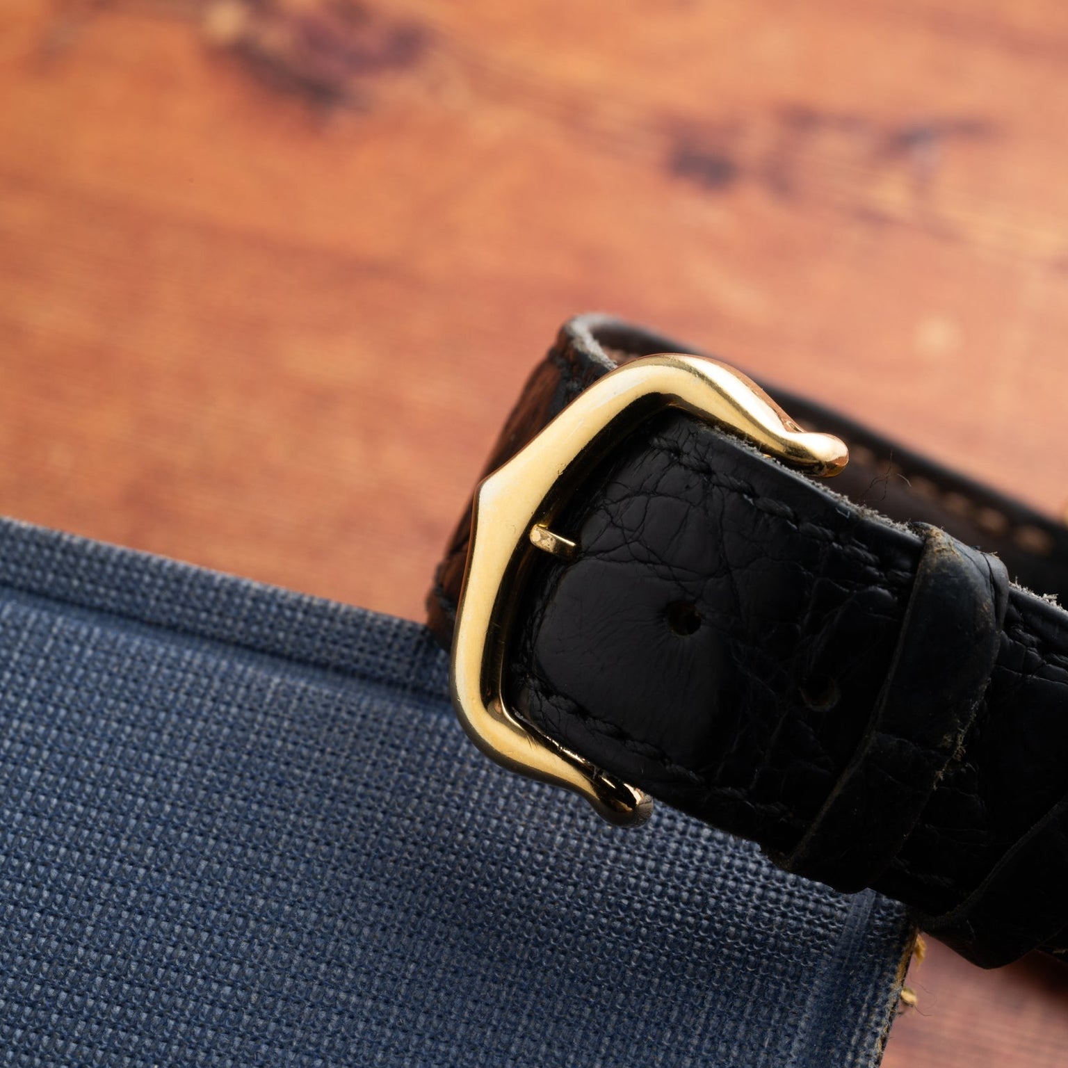 The Cartier Calandre black leather watch strap with a gold buckle is displayed on a blue fabric surface.