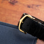 The Cartier Calandre black leather watch strap with a gold buckle is displayed on a blue fabric surface.