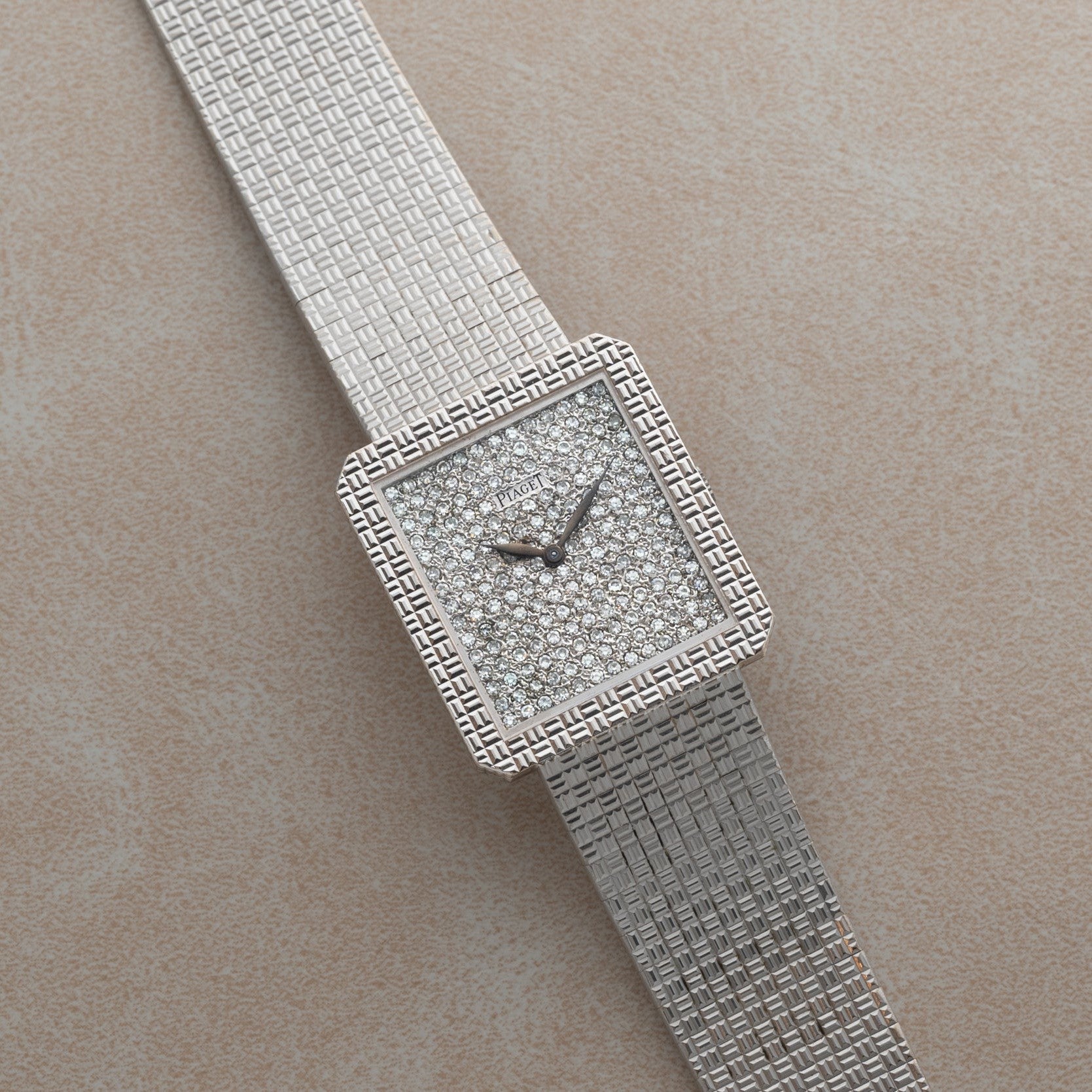 The Piaget Brick Diamond Dress Watch is a silver square luxury wristwatch with a diamond-encrusted face and textured metal band, displayed on a beige surface.