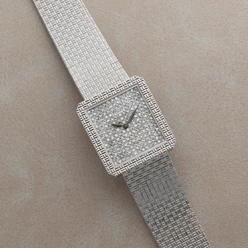 The Piaget Brick Diamond Dress Watch is a silver square luxury wristwatch with a diamond-encrusted face and textured metal band, displayed on a beige surface.