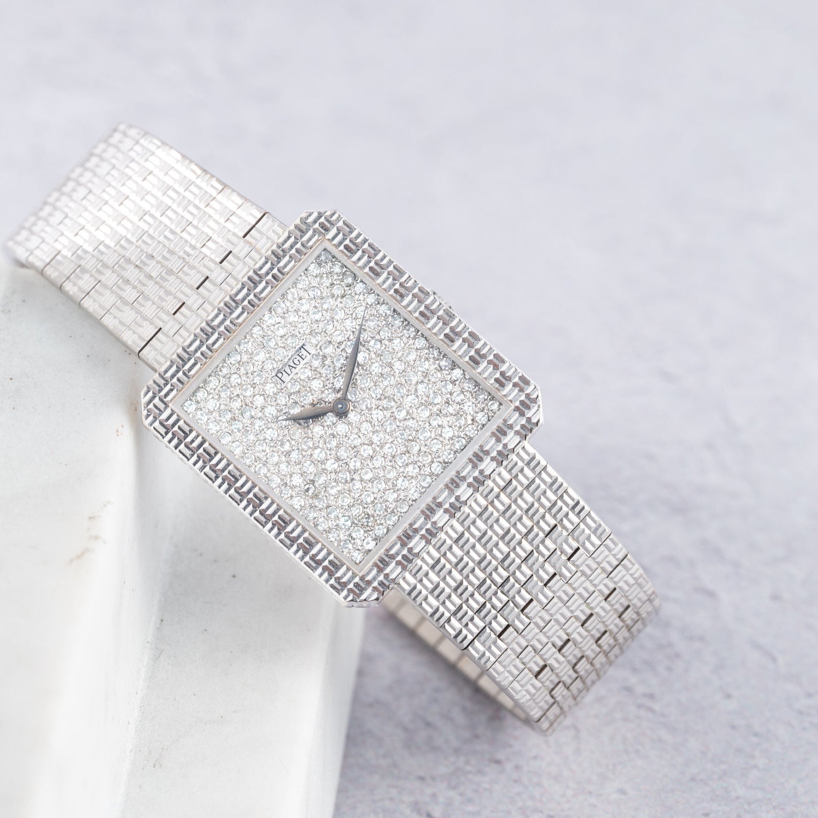 The Piaget Brick Diamond Dress Watch features a silver square face with a diamond-encrusted dial and band, elegantly displayed on a light marble surface.