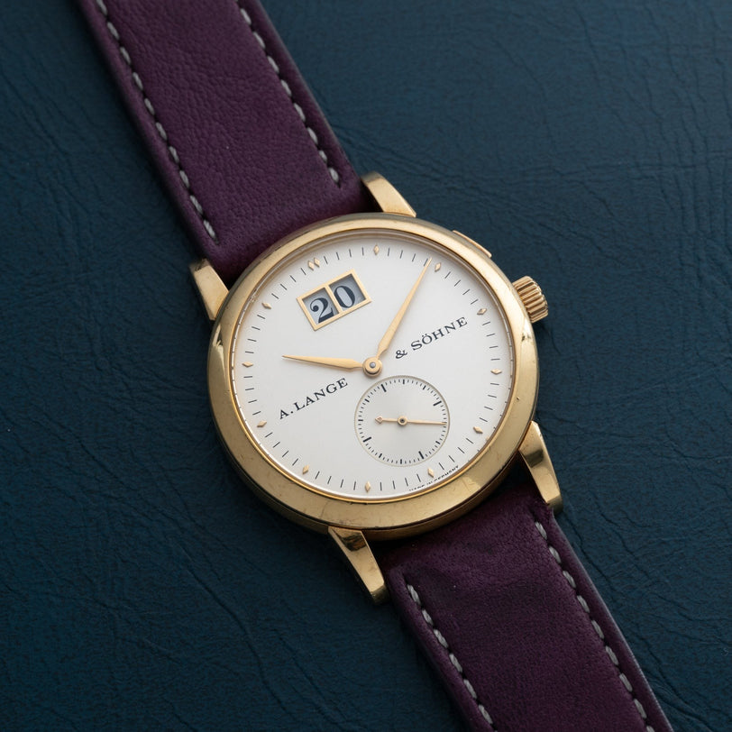 The Lange & Sohne Saxonia Outsize Date watch features a white dial, large date display, and purple strap, shown on a dark surface.