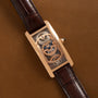 The Cartier Tank Cintreé Skeleton is a rectangular gold watch with a skeleton dial and brown leather strap, displayed on a brown leather surface.
